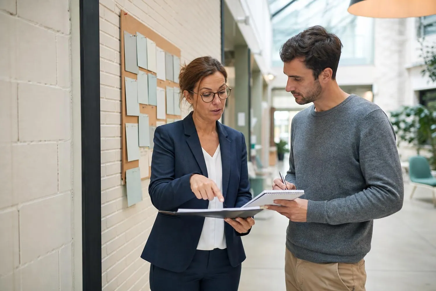Deux professionnels des relations entreprises font le point debout devant un tableau de suivi de campagne dans le hall clair d’un etablissement d’enseignement